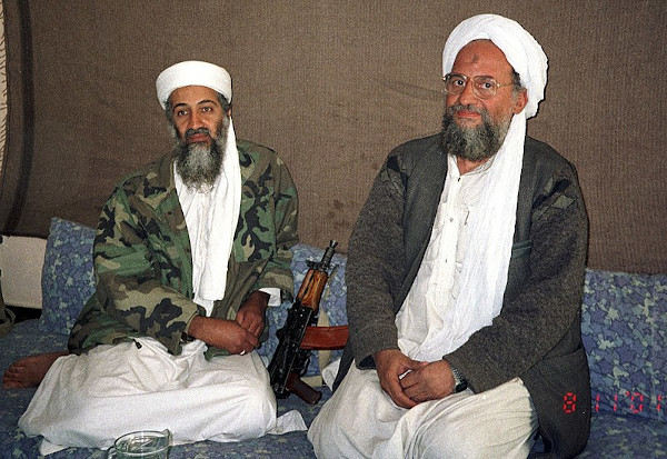 Osama bin Laden e Ayman al-Zawahiri sentados sobre tecido e almofadas, ao lado de uma arma de fogo.