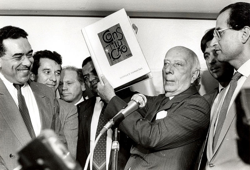 Ulysses GuimarÃ£es segurando a ConstituiÃ§Ã£o de 1988, um marco na histÃ³ria da redemocratizaÃ§Ã£o do Brasil.