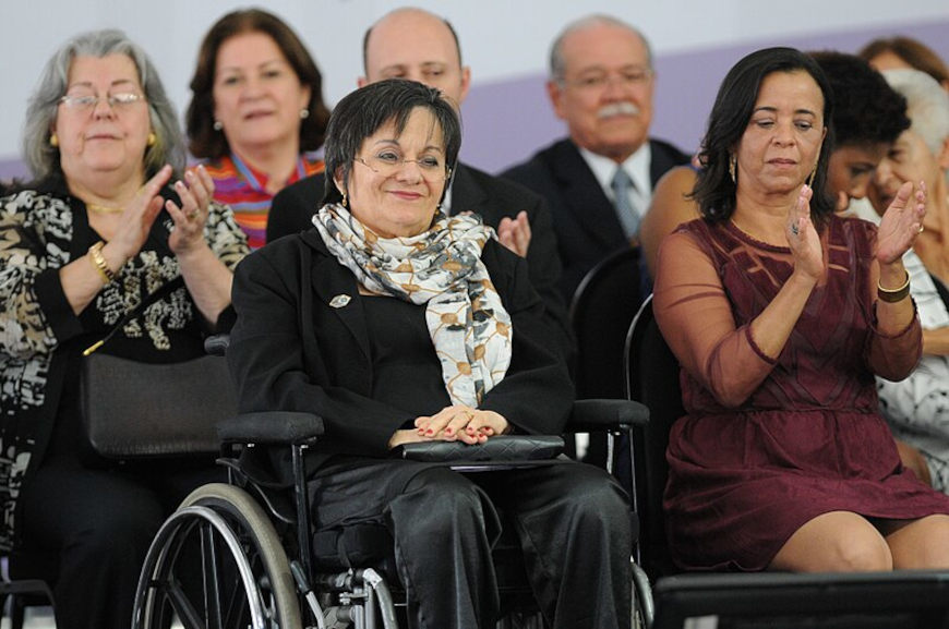 Maria da Penha sentada em sua cadeira de rodas, entre outras pessoas.