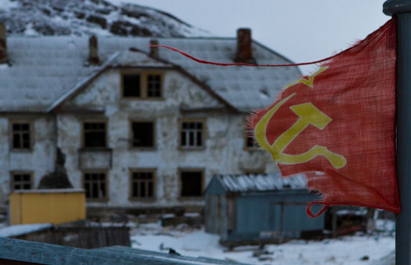 Bandeira da União Soviética rasgada em frente a uma casa em ruínas, em alusão aos efeitos da perestroika e da glasnost.